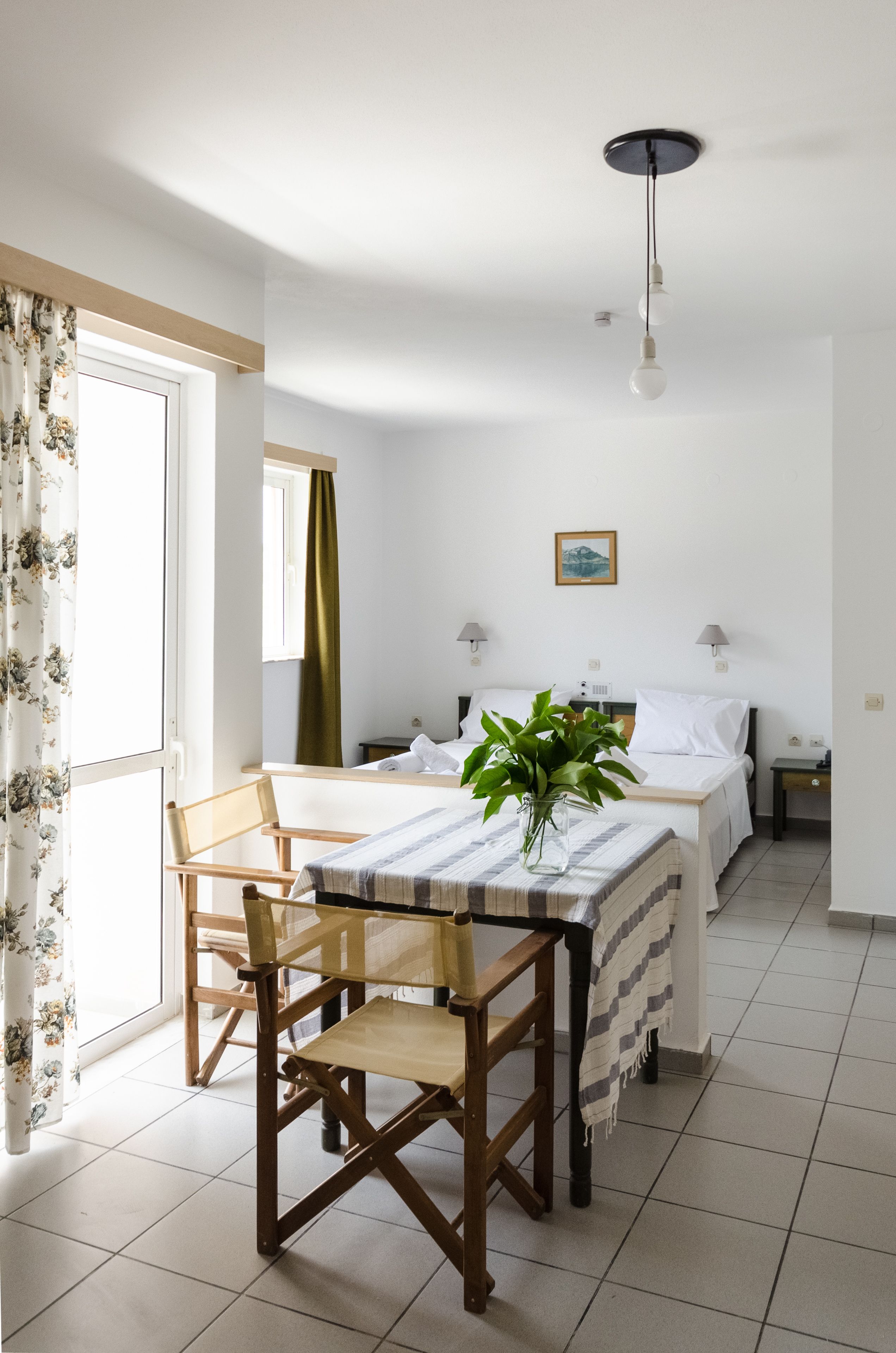 A room with a bed, bedside tables, a desk with a chair, a hanging light fixture, and a window with curtains.