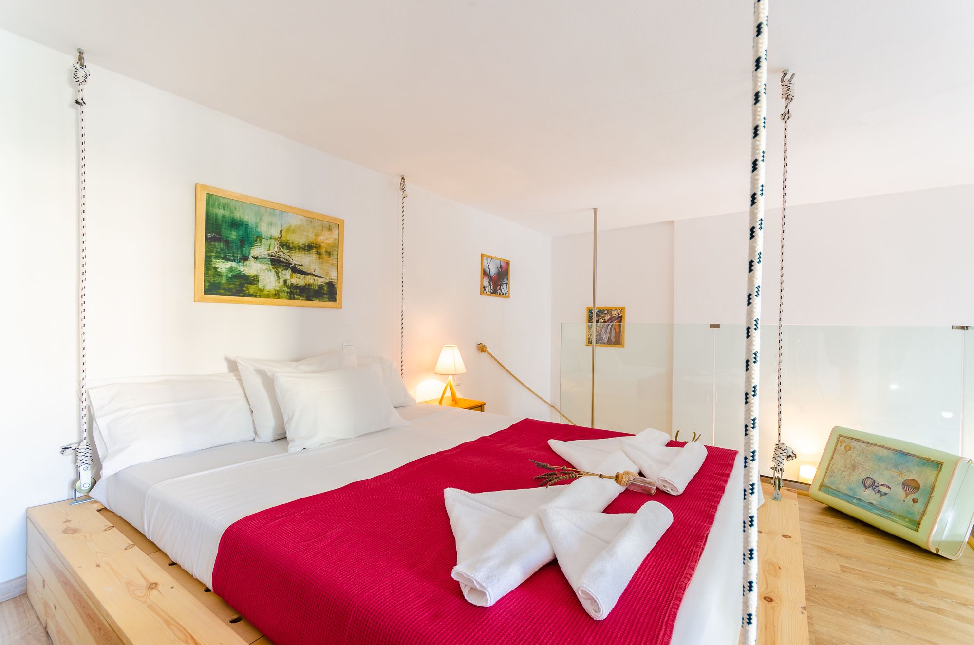 Un dormitorio con cama, una manta roja, toallas blancas, una mesita de noche, una lámpara y un cuadro en la pared.