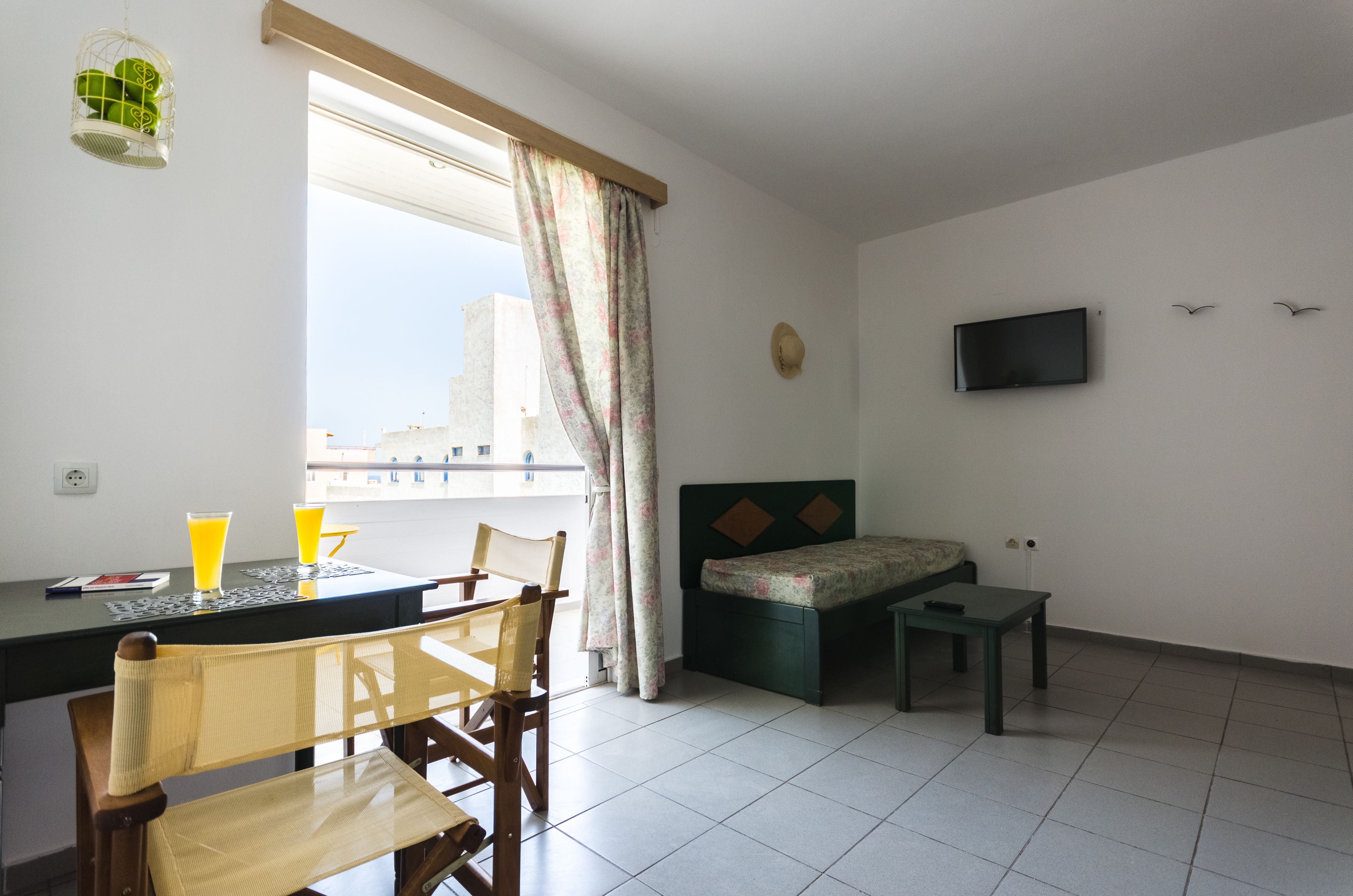 A small, bright room with a bed, a TV, a table with chairs, and a window with curtains.
