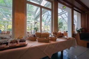 A breakfast buffet table with various food items and baskets, set in a room with large windows and a view of greenery outside. - Albhotel Fortuna (Riederich)