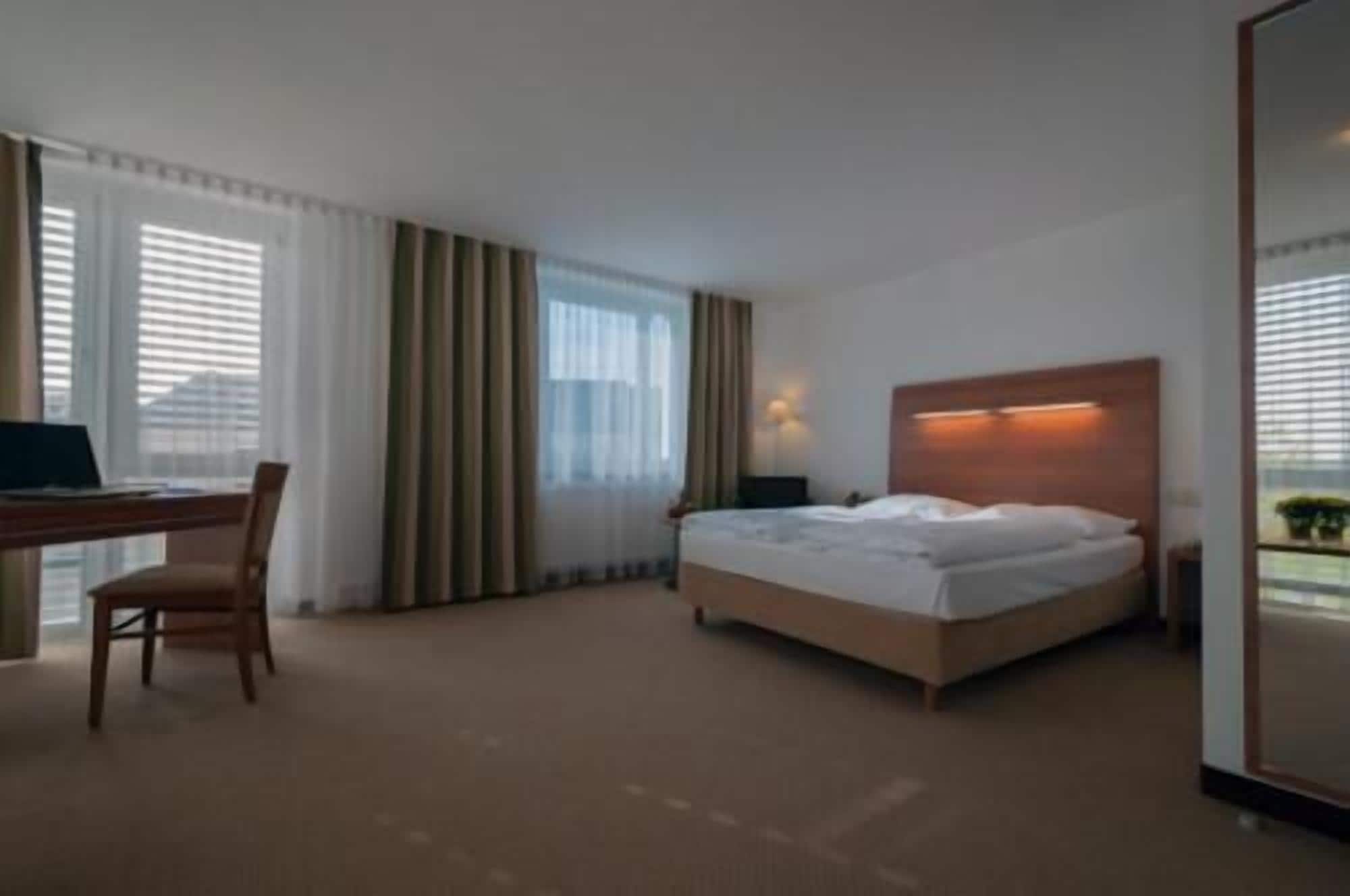 Habitación de hotel con cama, escritorio y silla. Se aprecia una ventana con cortinas y una lámpara en la pared.