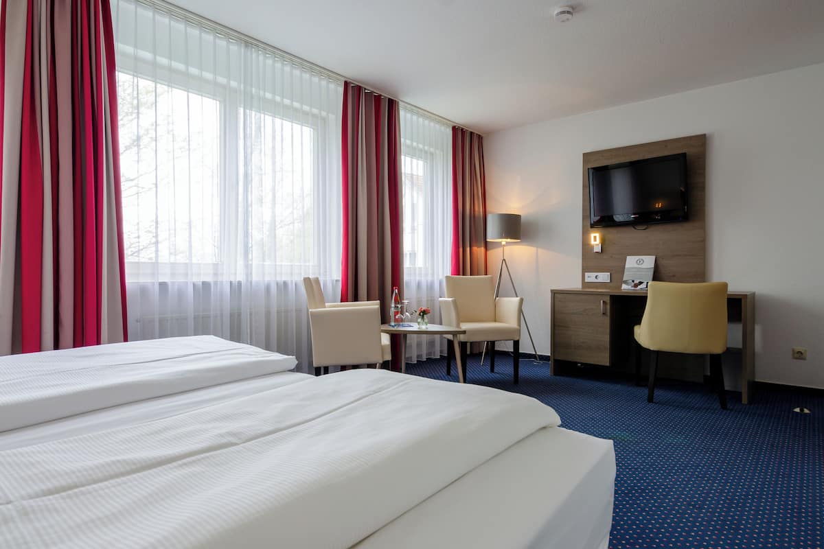 A hotel room with a large bed, a desk, two chairs, a TV mounted on the wall, and a window with curtains.