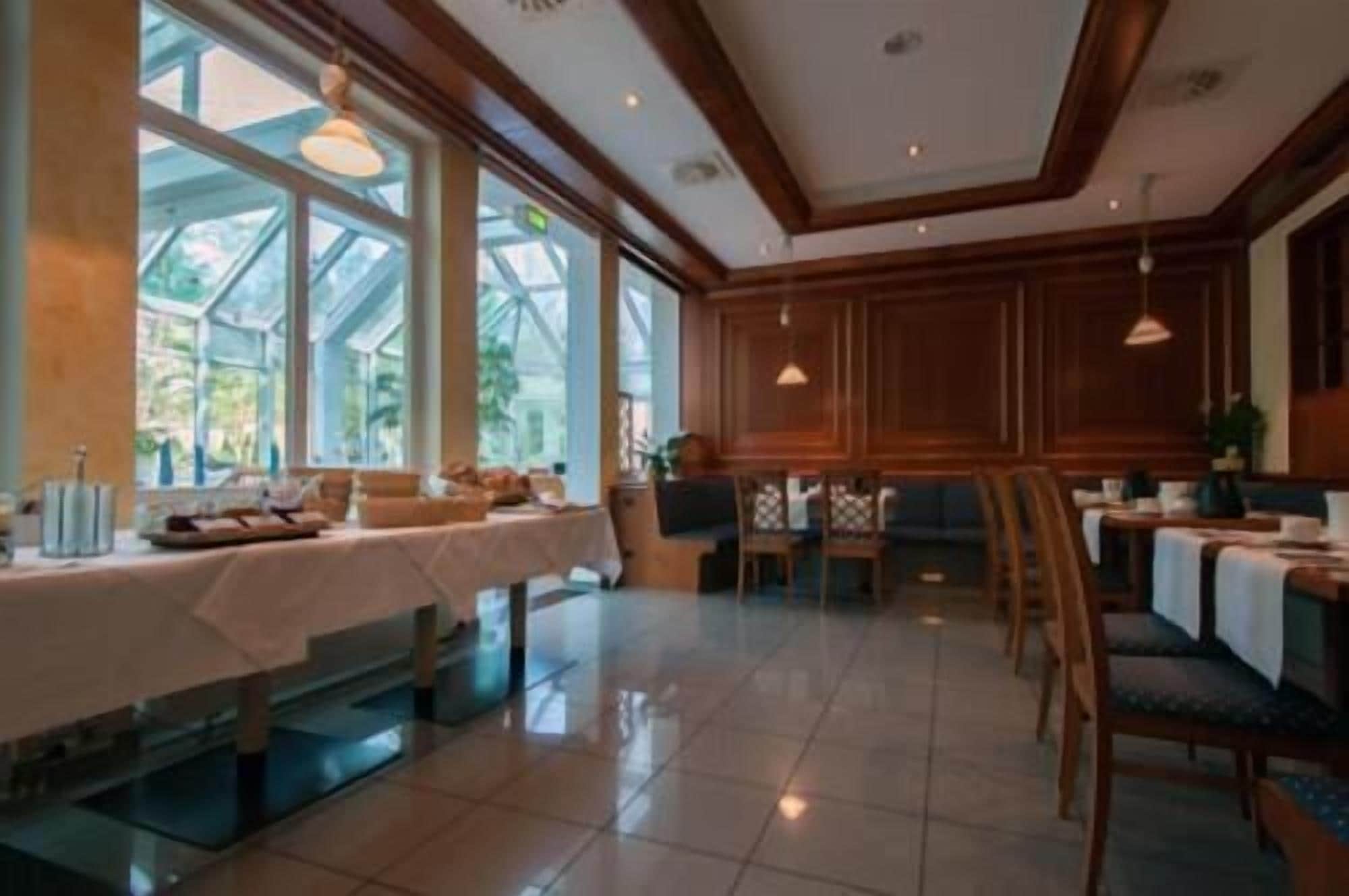 Un comedor amplio con techos altos, ventanas grandes y paneles de madera. Las mesas están preparadas para comer, y las sillas dispuestas alrededor.
