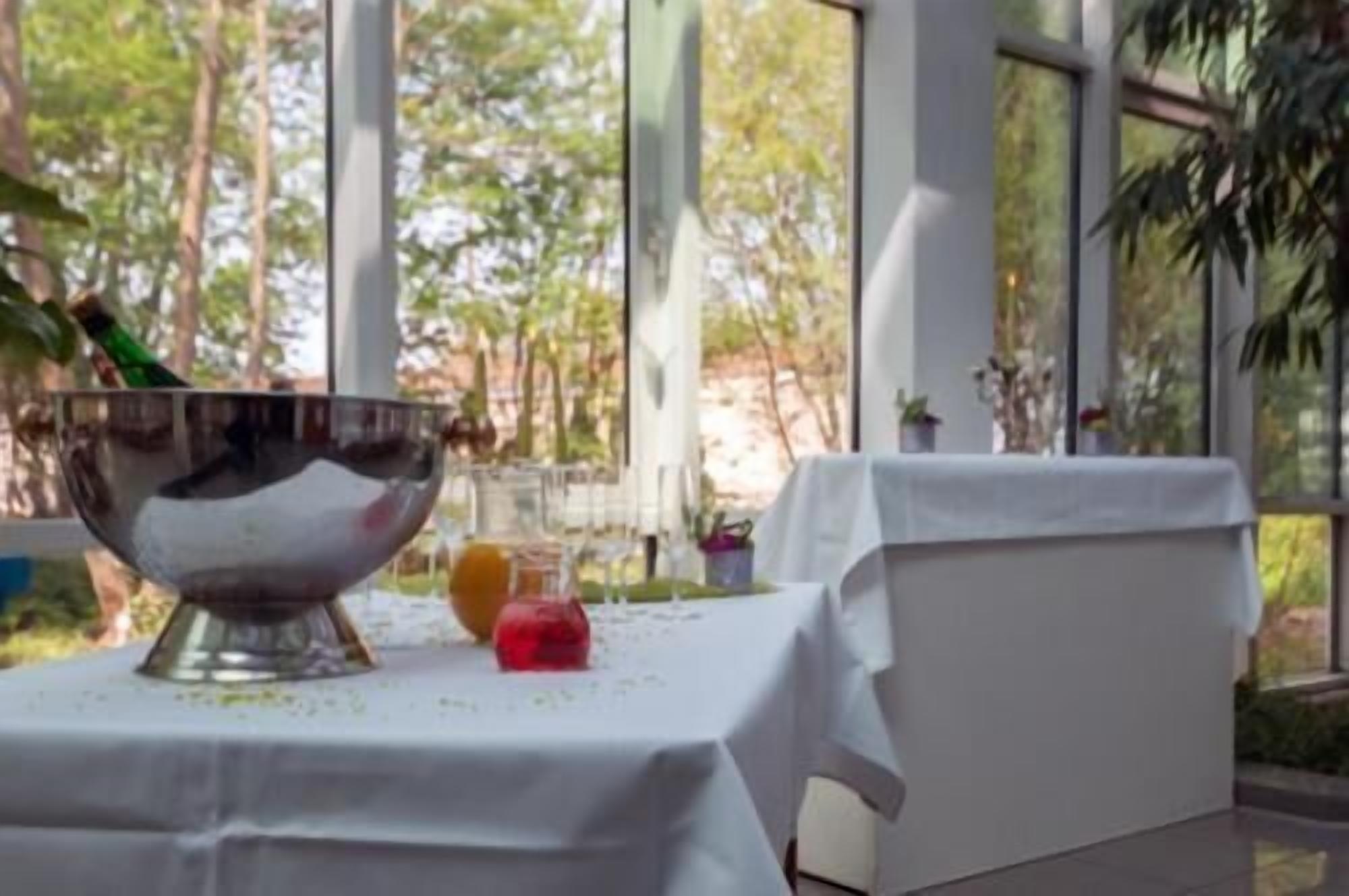Una mesa con mantel blanco, un cuenco metálico y un objeto rojo, frente a grandes ventanales con vistas a árboles.