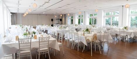 A spacious banquet hall with multiple round tables covered in white tablecloths, arranged in rows.