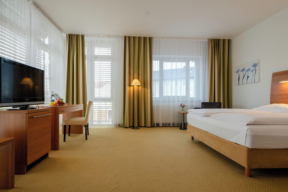 A hotel room with a bed, a desk with a TV, a chair, a lamp, and a small table with flowers.