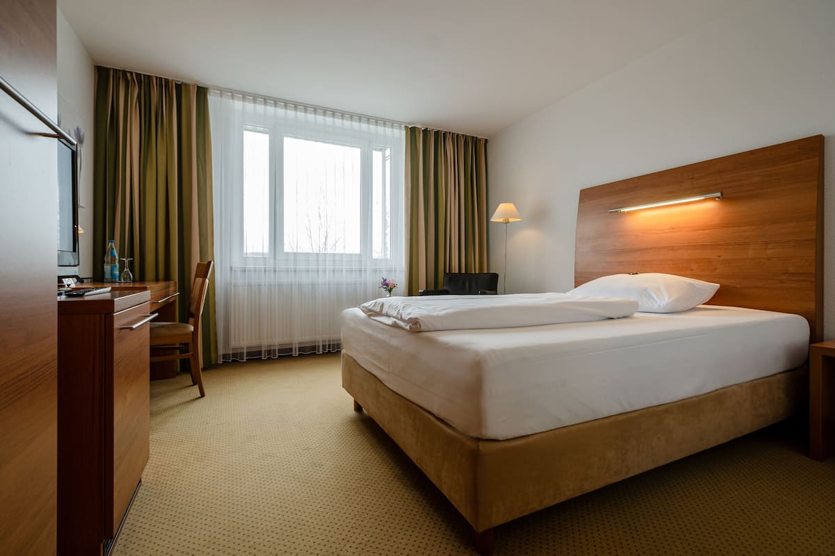 A hotel room with a bed, a desk, a chair, a lamp, and a window with curtains.