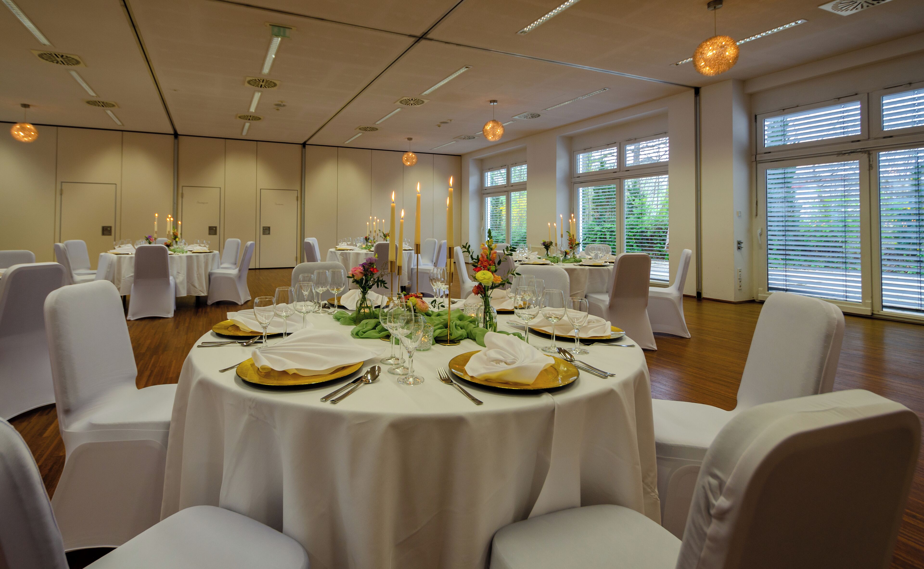 Un salón de banquetes con mesas redondas dispuestas para comer, con manteles blancos, platos base dorados y centros de mesa con velas.