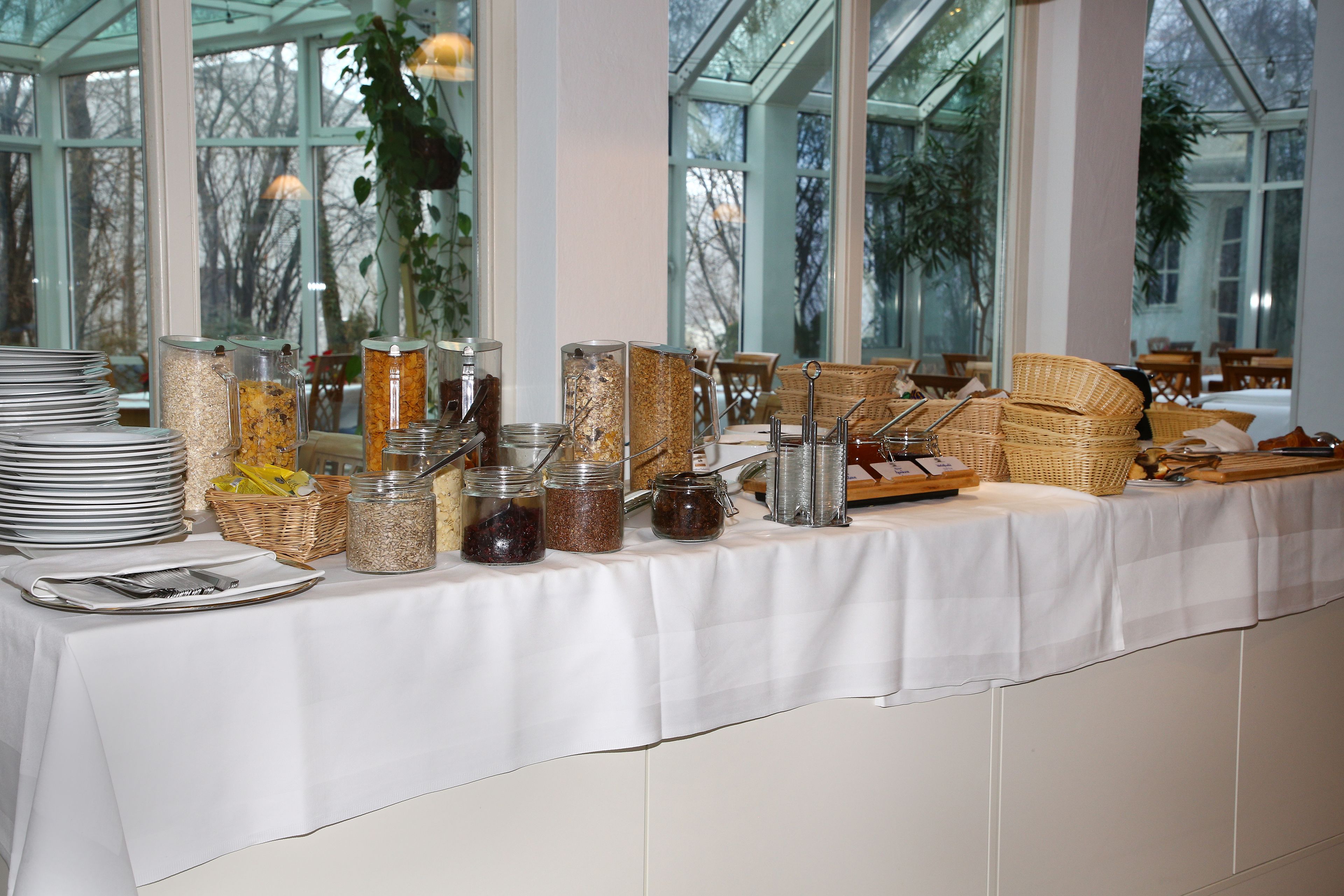 A buffet table with various food containers and utensils, set in a room with large windows and a view of trees.
