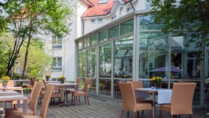 An outdoor café with wicker chairs and tables, large glass windows, and a building with a red-tiled roof in the background. - Albhotel Fortuna (Riederich)