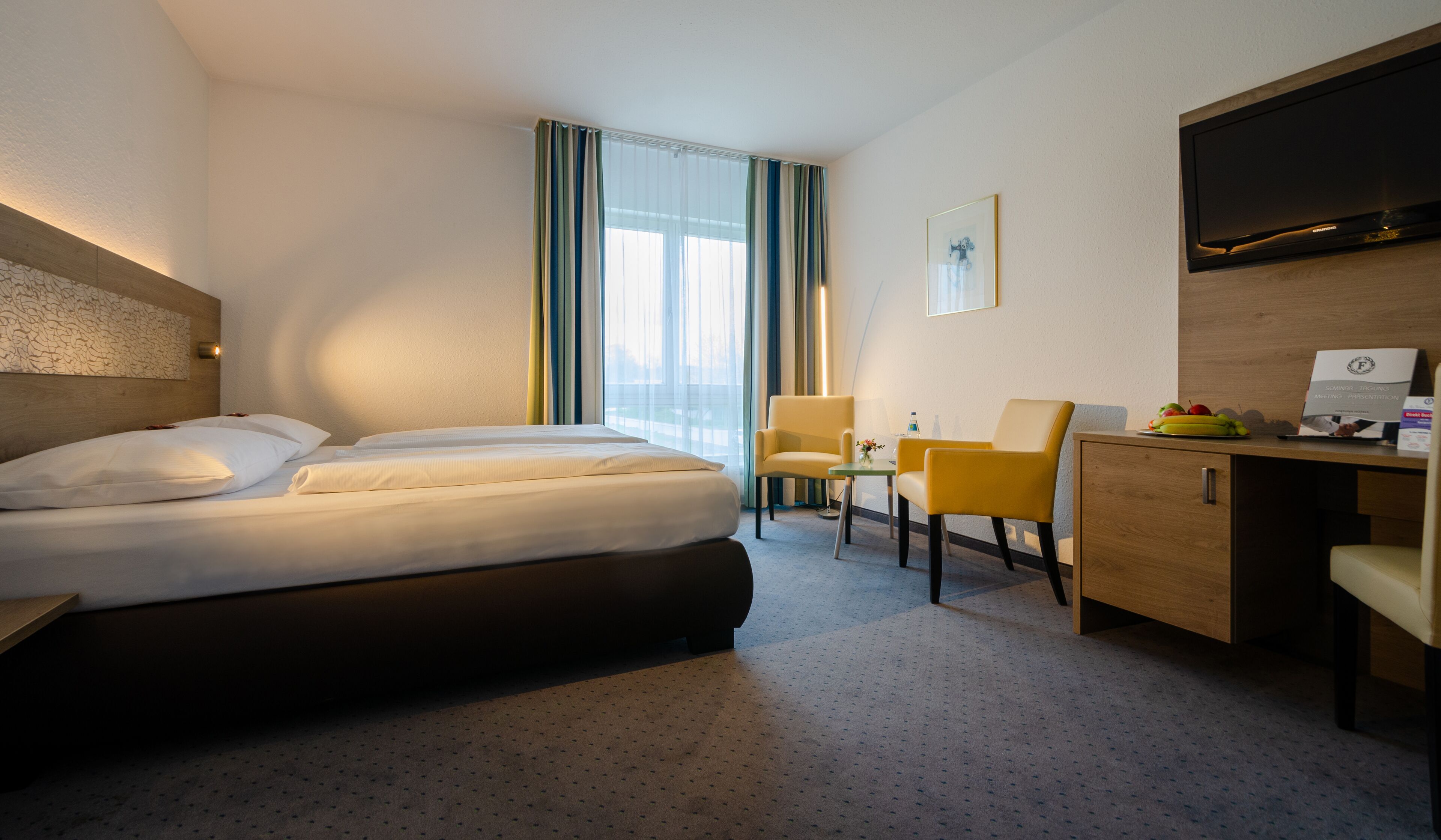 Ein Hotelzimmer mit Bett, Schreibtisch, Stuhl, Fernseher und einem Fenster mit Vorhängen.