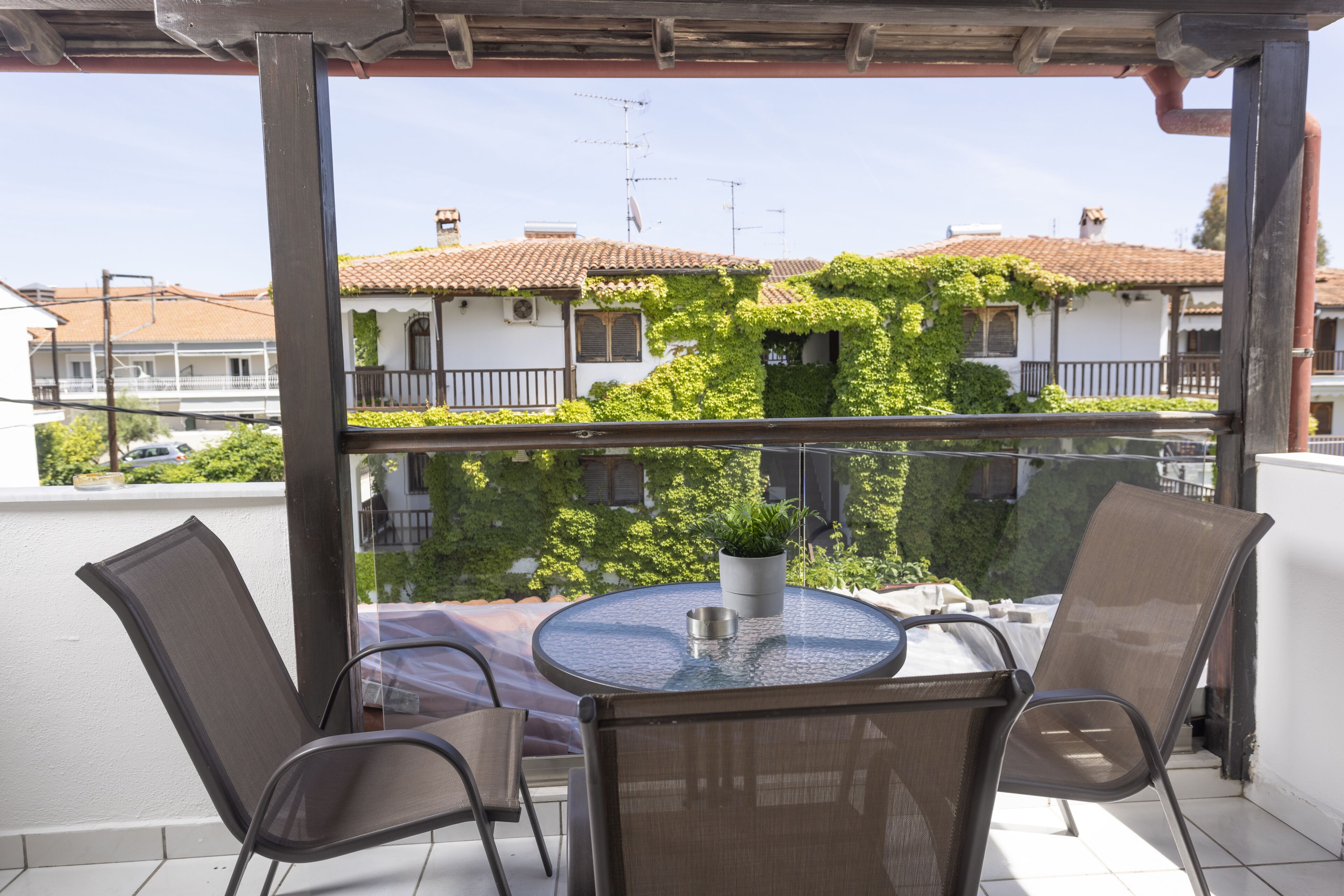 A balcony with a round glass table and two chairs, overlooking a residential area with houses and greenery.