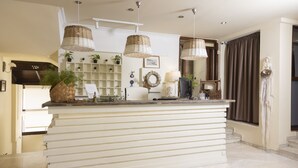 A reception area with a white reception desk, wicker pendant lights, a potted plant, and a decorative wreath on the wall. - Hotel Magdalena (Kassandra)