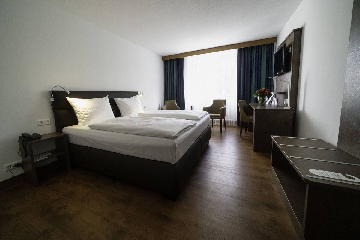 A hotel room with a large bed, a desk, a chair, a TV, and a window with curtains.