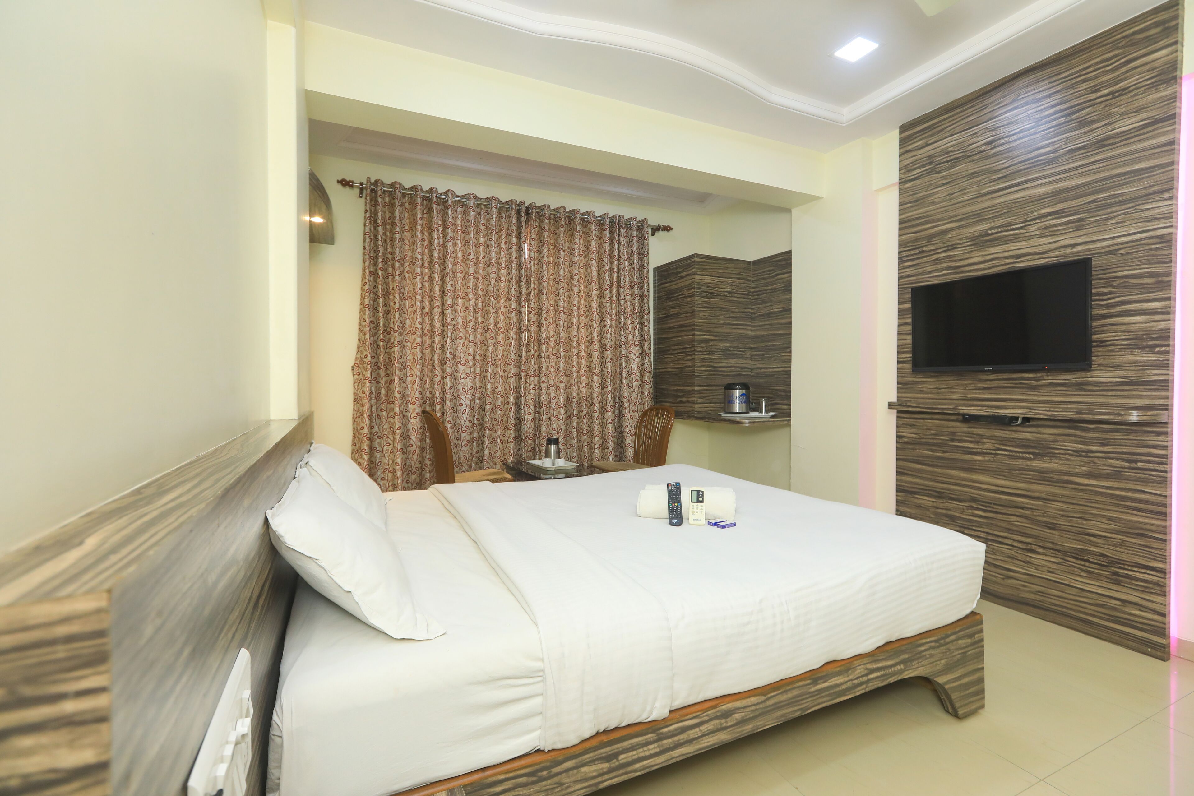 A hotel room with a bed, a TV mounted on the wall, a curtain, and a bedside table with a bottle and remote controls.