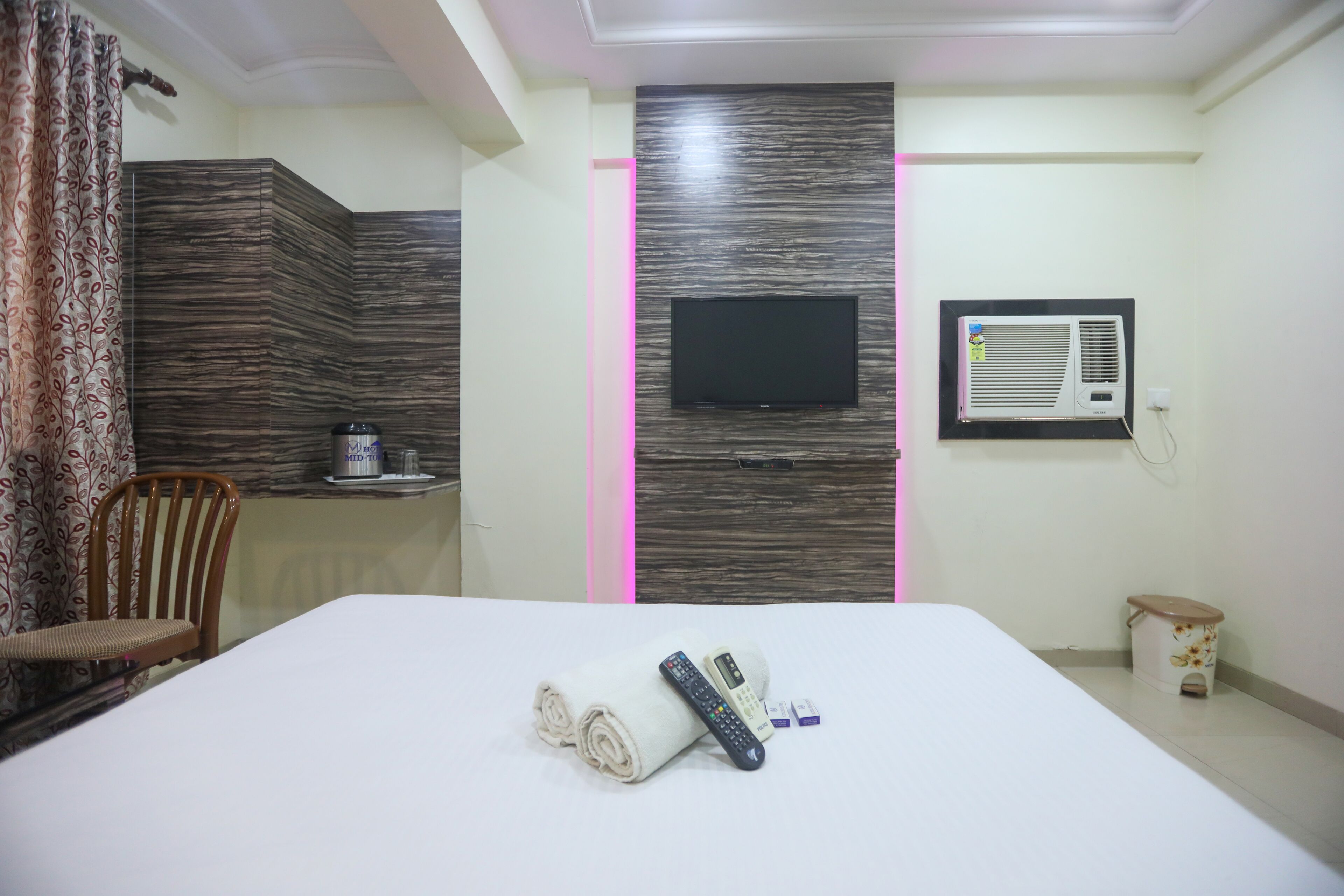 A hotel room with a bed, a TV mounted on the wall, a small table with a kettle, a chair, and a trash bin.