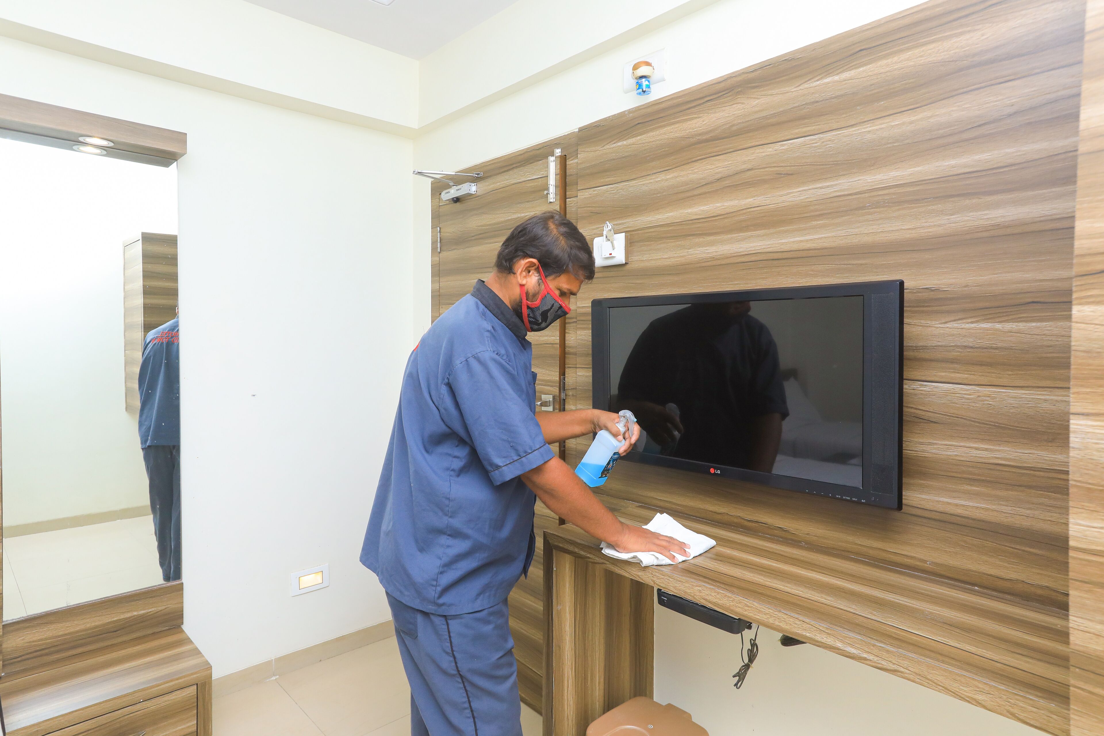 A person cleaning a wall-mounted television in a room with a wooden panel design and a mirror reflecting part of the room.