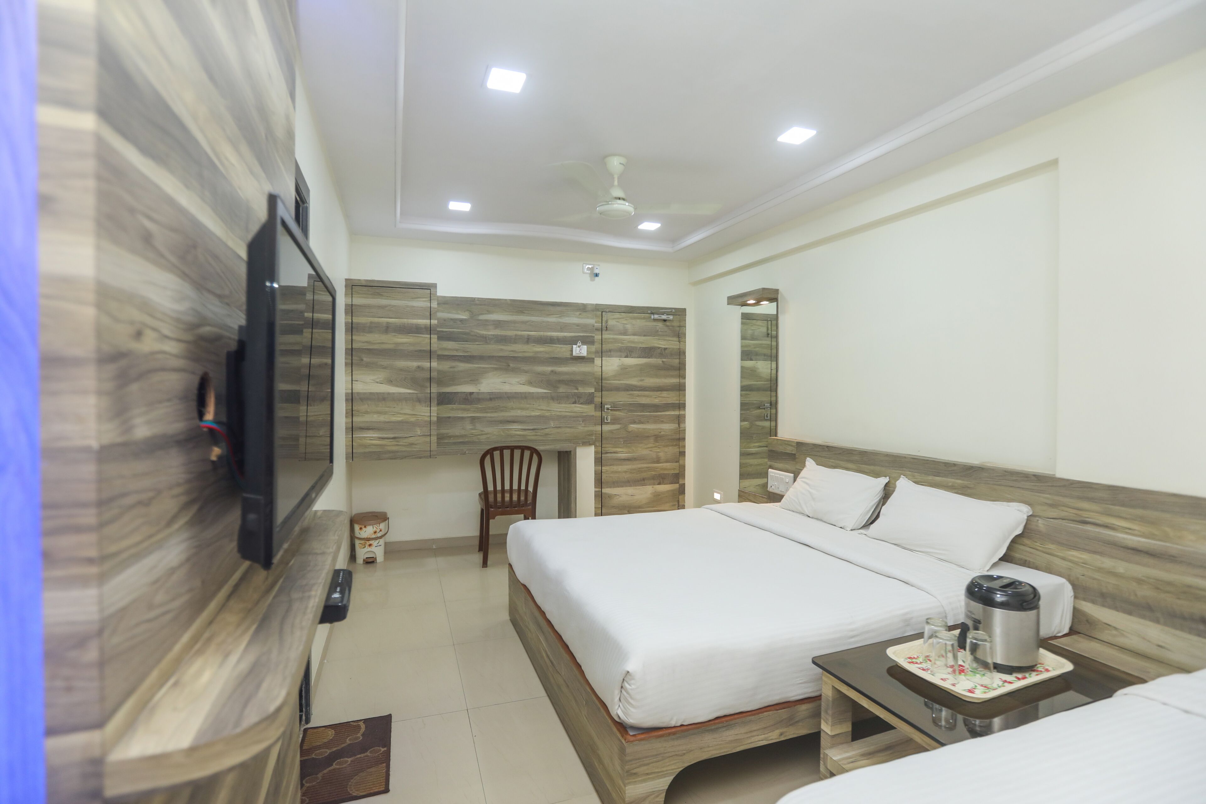 A hotel room with a large bed, a bedside table with a kettle, a chair, and a TV mounted on the wall.