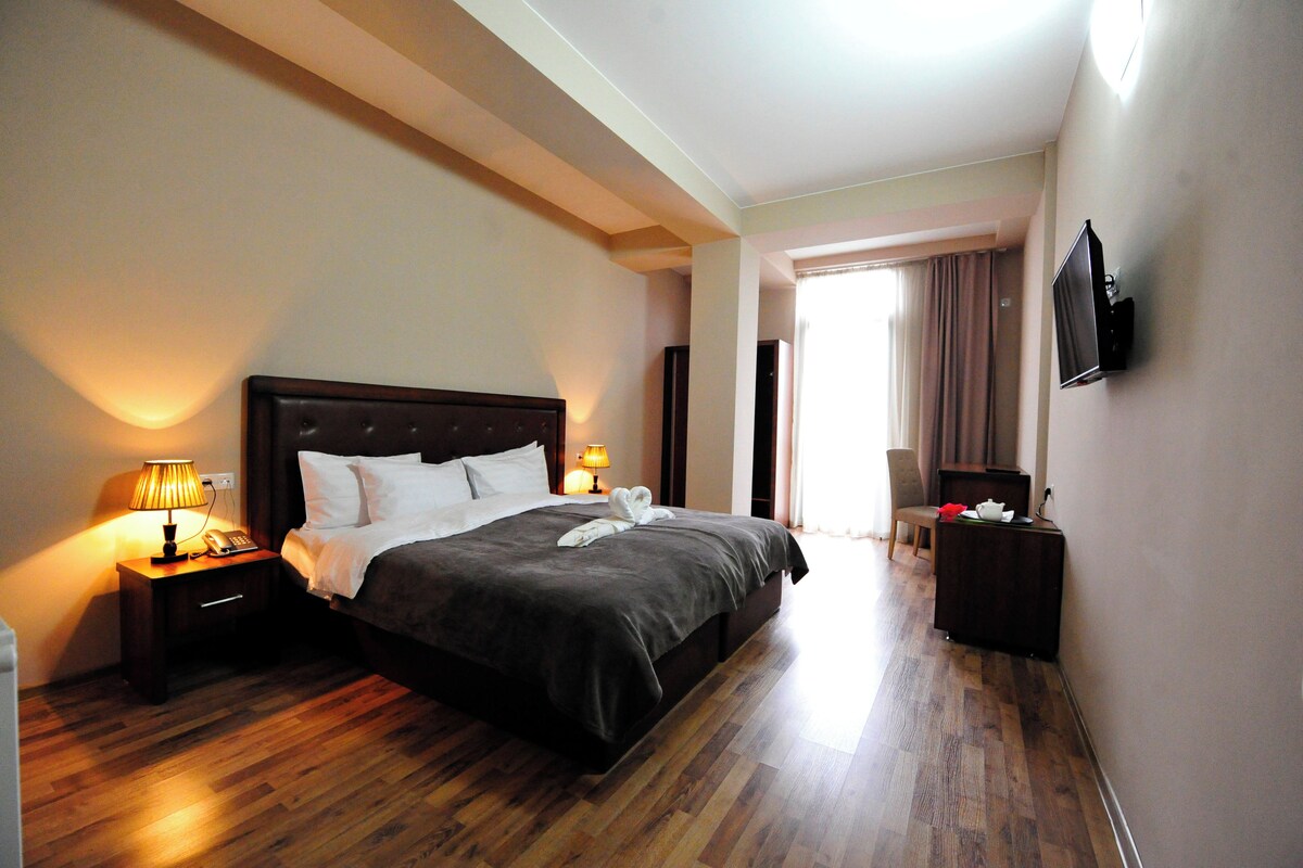 A hotel room with a large bed, bedside lamps, a flat-screen TV mounted on the wall, and a wooden floor.