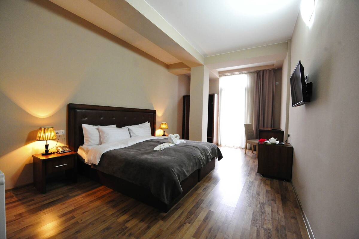 A hotel room with a large bed, bedside lamps, a flat-screen TV mounted on the wall, and a wooden floor.