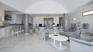 A spacious lobby with a high ceiling, marble flooring, and a central seating area with sofas and chairs. There is a reception desk with a person behind it, and a bar area with shelves of bottles. - Aperanto Suites (Santorini)