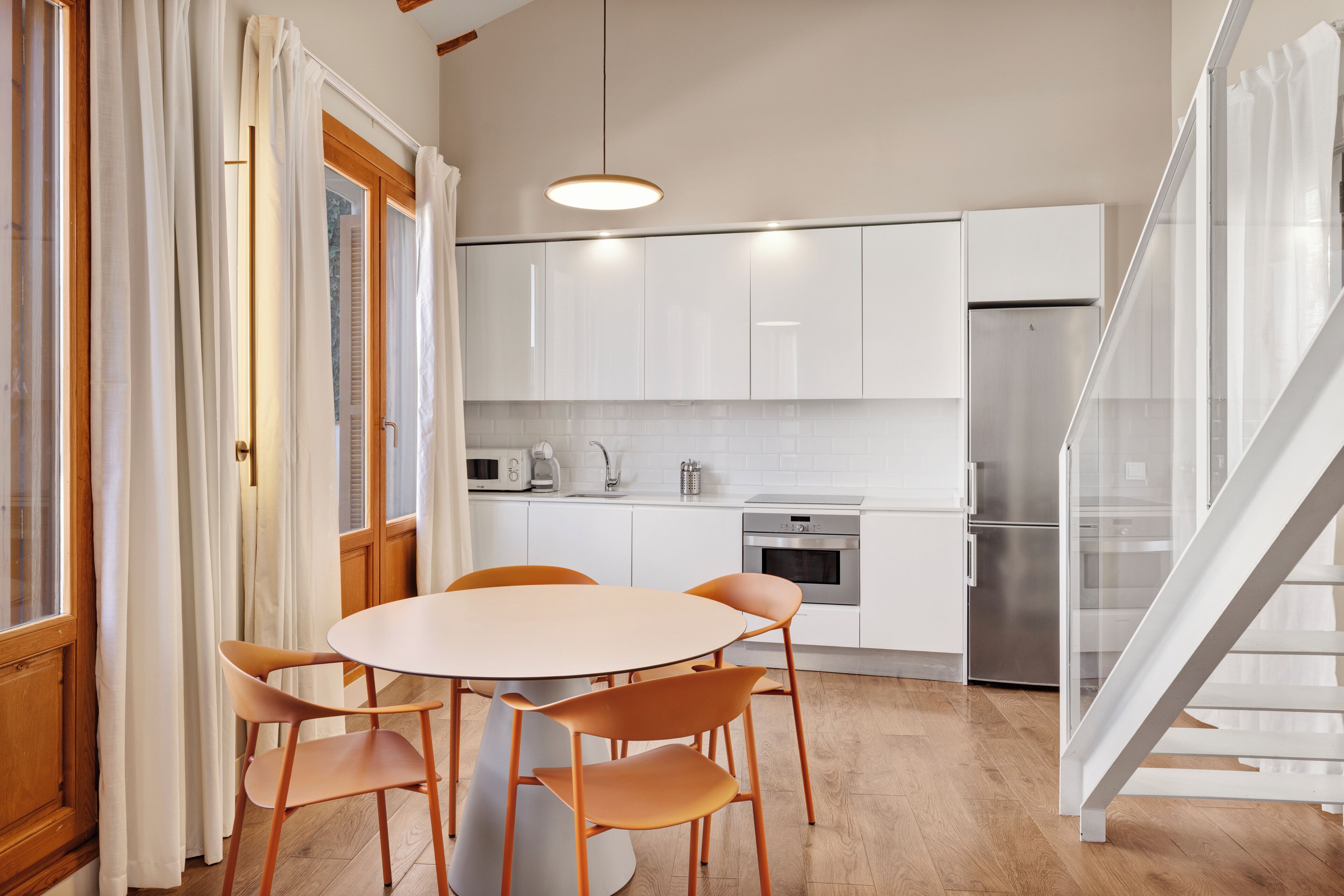 Una cocina moderna con armarios blancos, un refrigerador de acero inoxidable, una mesa de comedor con sillas y una escalera.