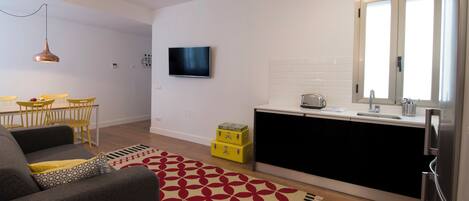 Un espacio habitable compacto con sofá, mesa de comedor, sillas, una zona de cocina con fregadero y armarios, y un televisor montado en la pared.