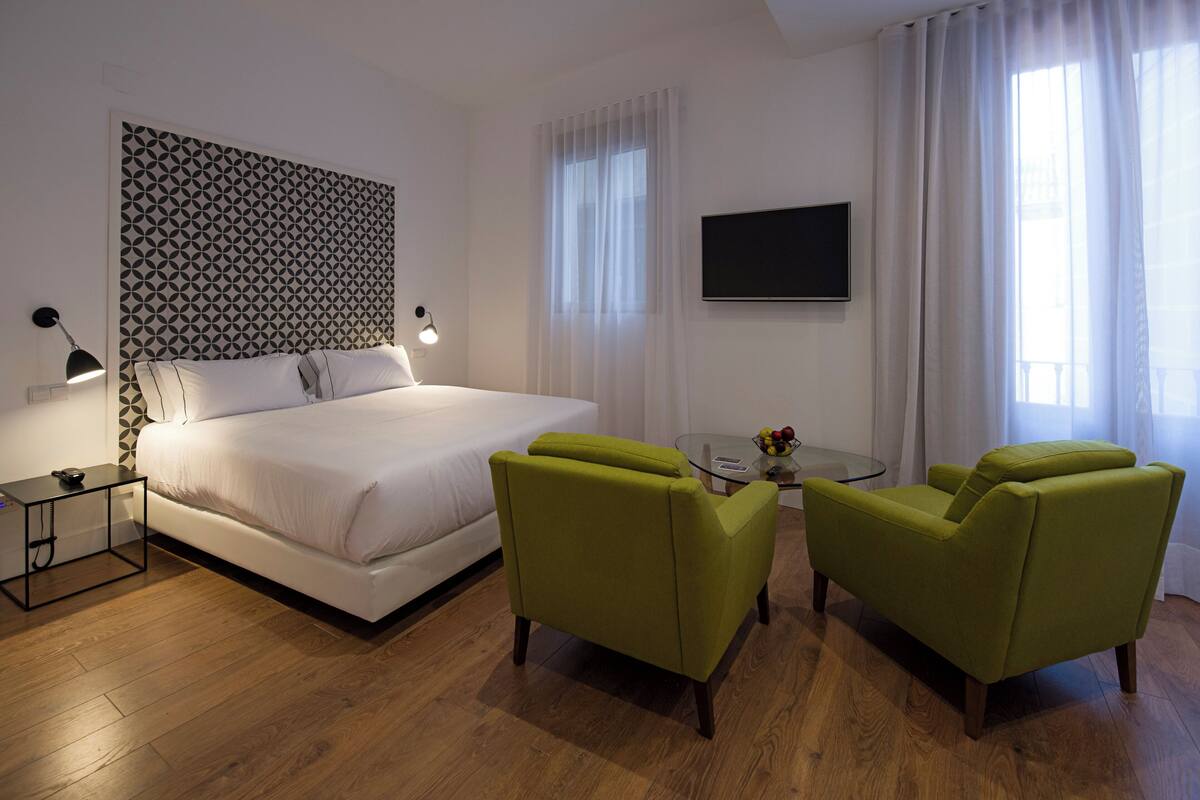 Una habitación de hotel con una cama, un sillón verde, una mesa de centro de vidrio, un televisor y un cabecero estampado.