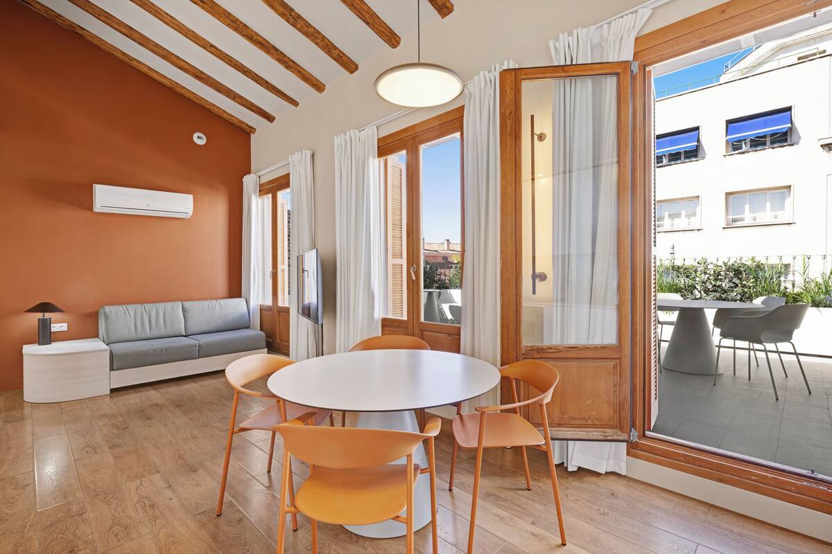 Una habitación con sofá, una mesa redonda con sillas, televisor y ventana con cortinas.