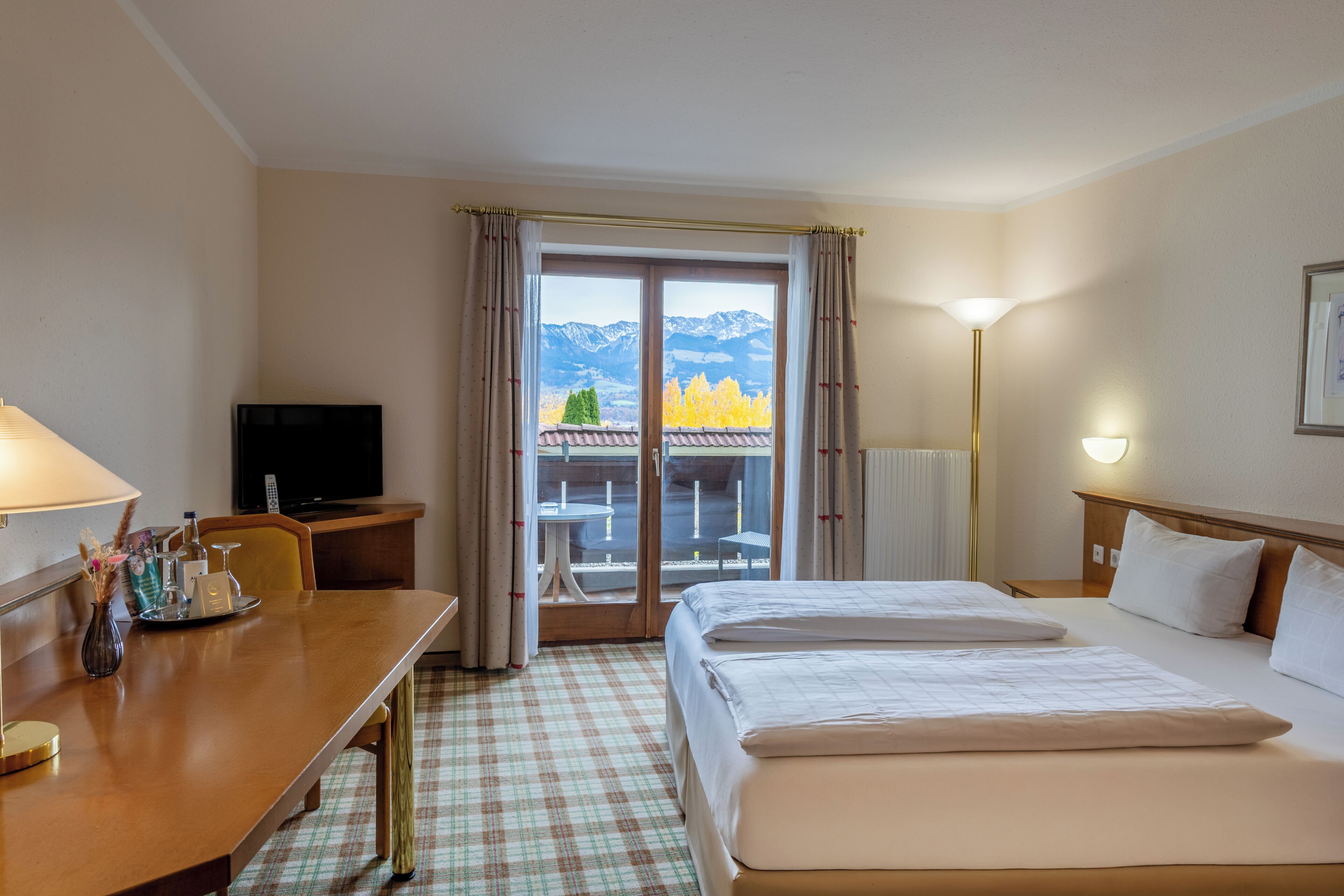 A hotel room with a bed, a desk with a lamp, a television, and a view of mountains through the window.