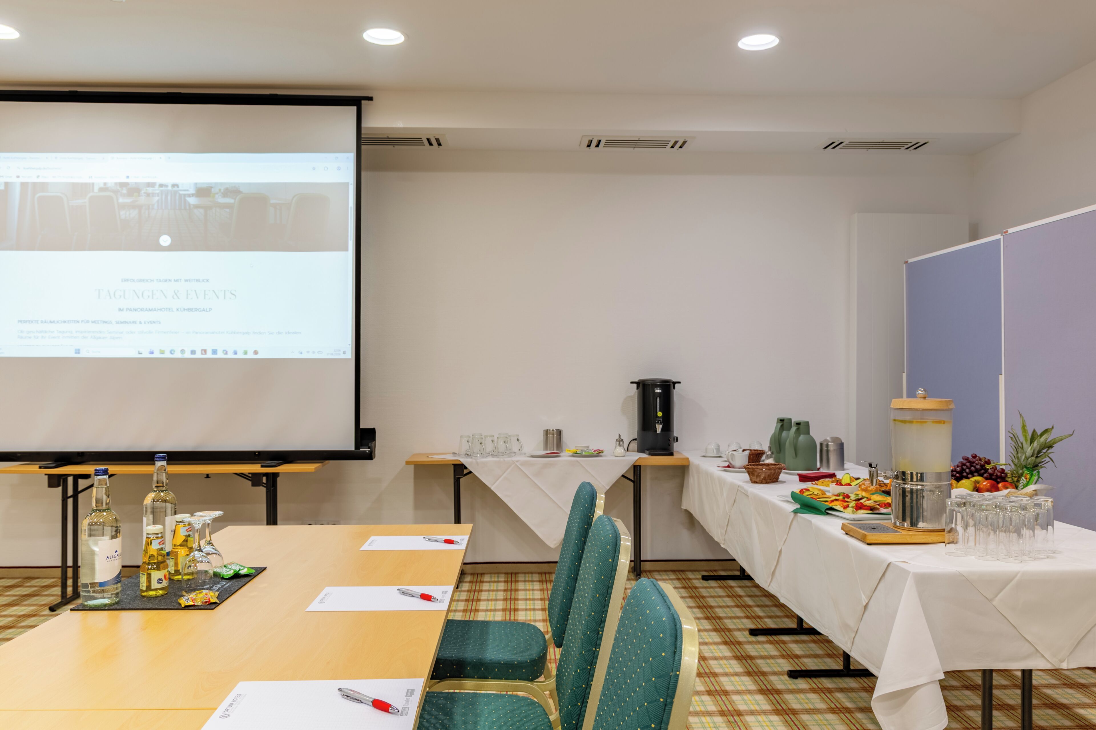 A conference room with a long table, chairs, a projector screen, and a buffet table with food and drinks.