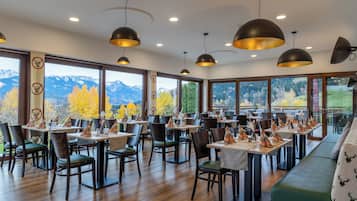 Un restaurante con varias mesas y sillas, amplias ventanas con vistas a la montaña e iluminación colgante.