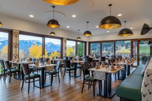 Un restaurante con varias mesas y sillas, amplias ventanas con vistas a la montaña e iluminación colgante.
