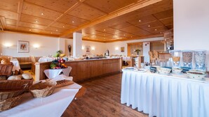 A spacious dining area with wooden ceiling beams, a buffet table with various food items, and a seating area with a bench and baskets. - Hotel Kuehbergalp (Blaichach)