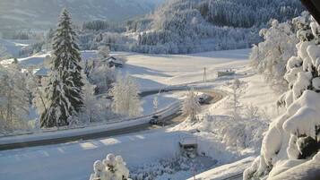 A snowy landscape with a road curving through a forested area, a small cabin, and a mountain range in the background.