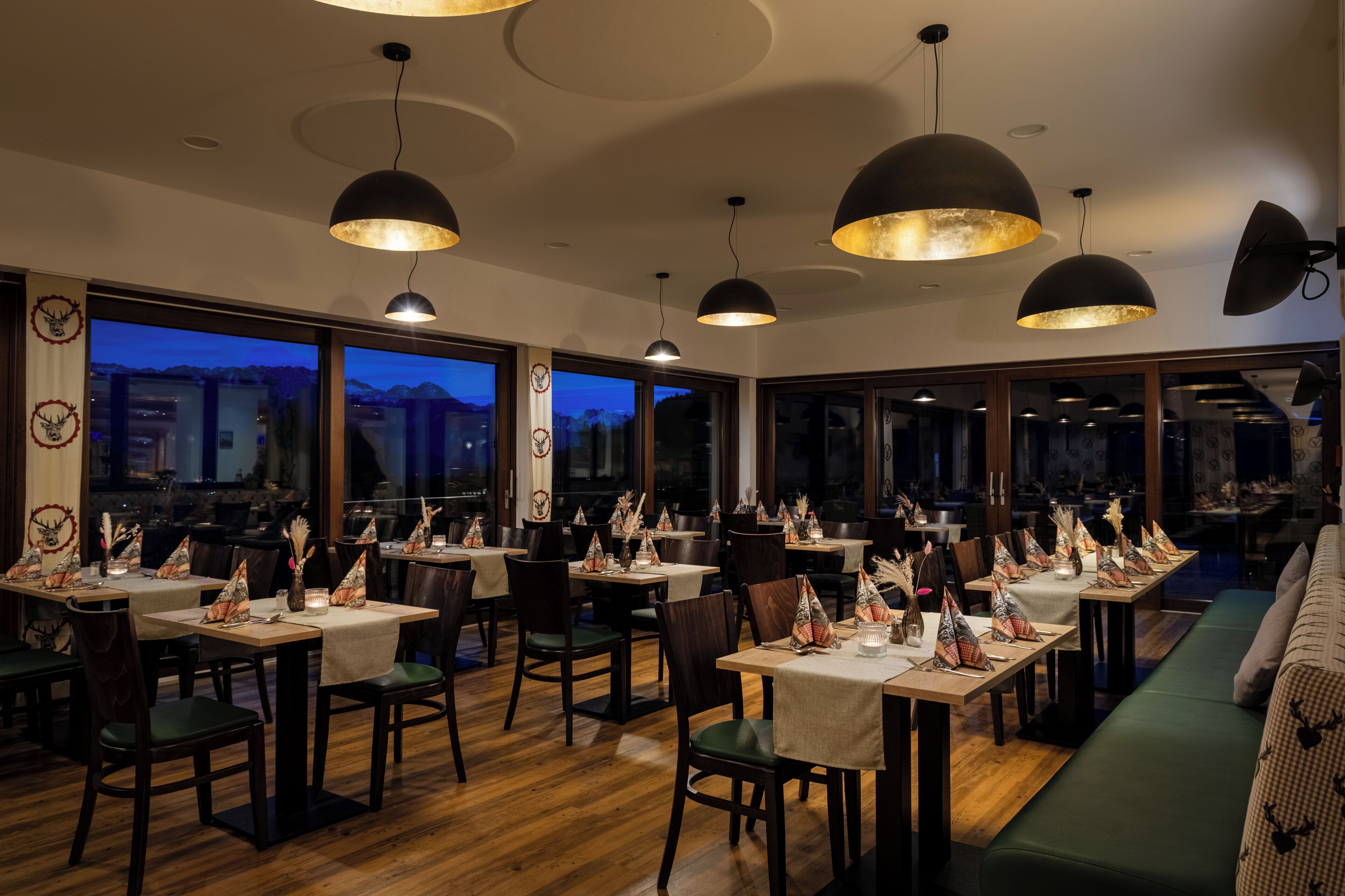 Restaurante com várias mesas preparadas para refeições, grandes janelas e iluminação pendente.