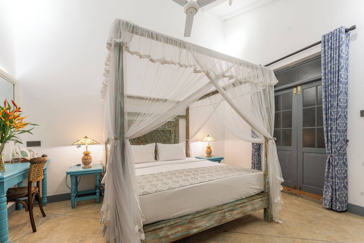 A four-poster bed with a canopy, a small table with a lamp, a chair, and a door with patterned curtains.