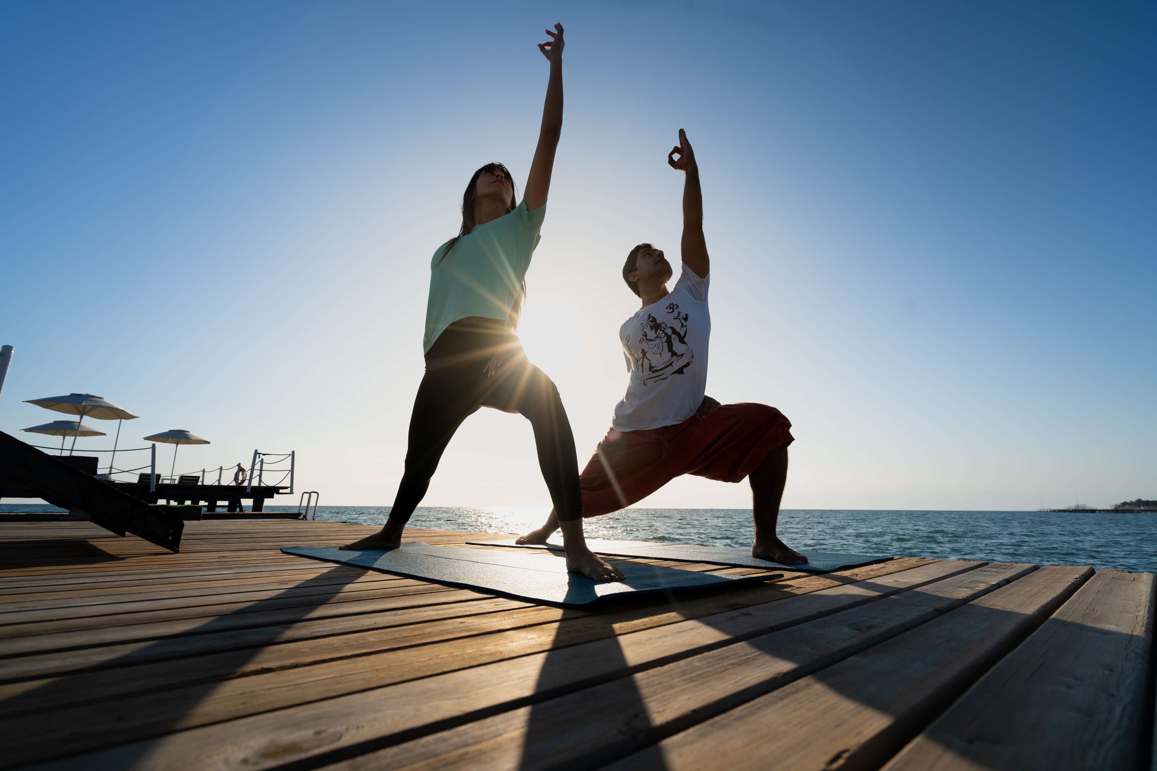 Two people practicing yoga on a wooden deck by the sea.