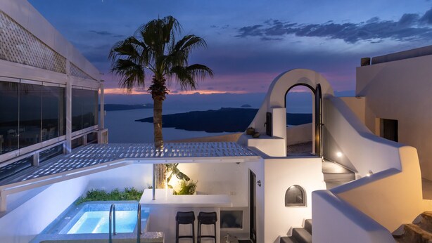 A modern outdoor pool area with a view of the sea, a palm tree, and a white building with a unique architectural design. - Opera Mansion (Santorini)
