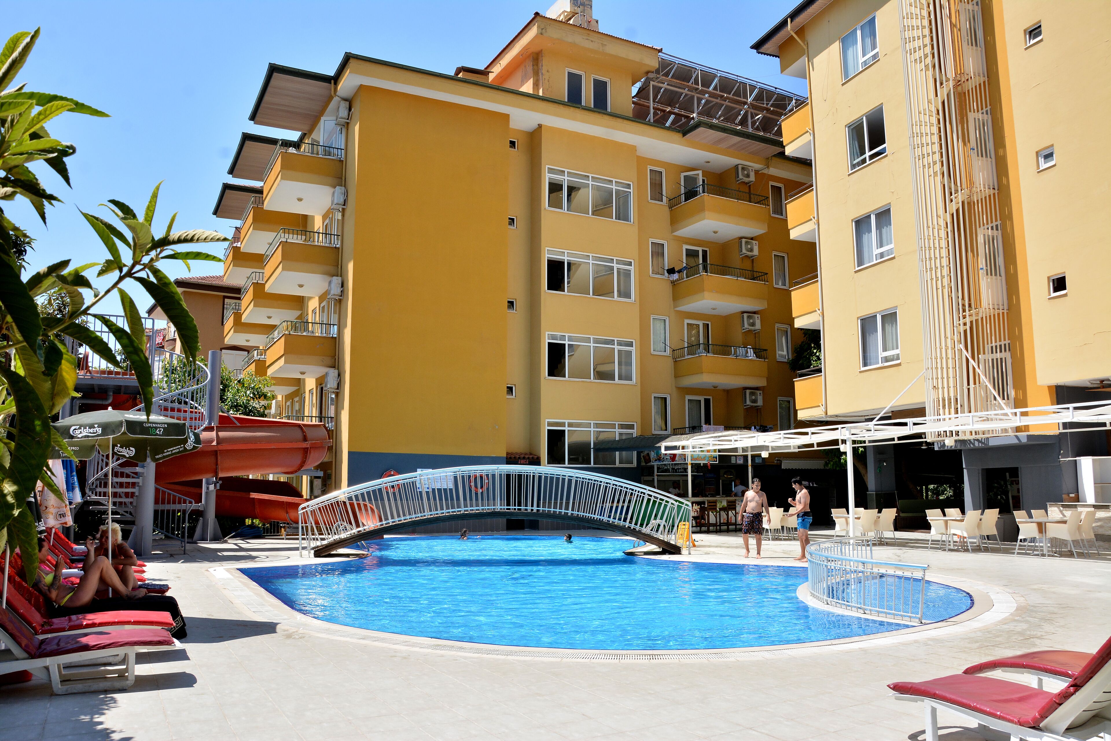 En swimmingpool omgivet af gule bygninger med balkoner, liggestole og en vandrutsjebane.