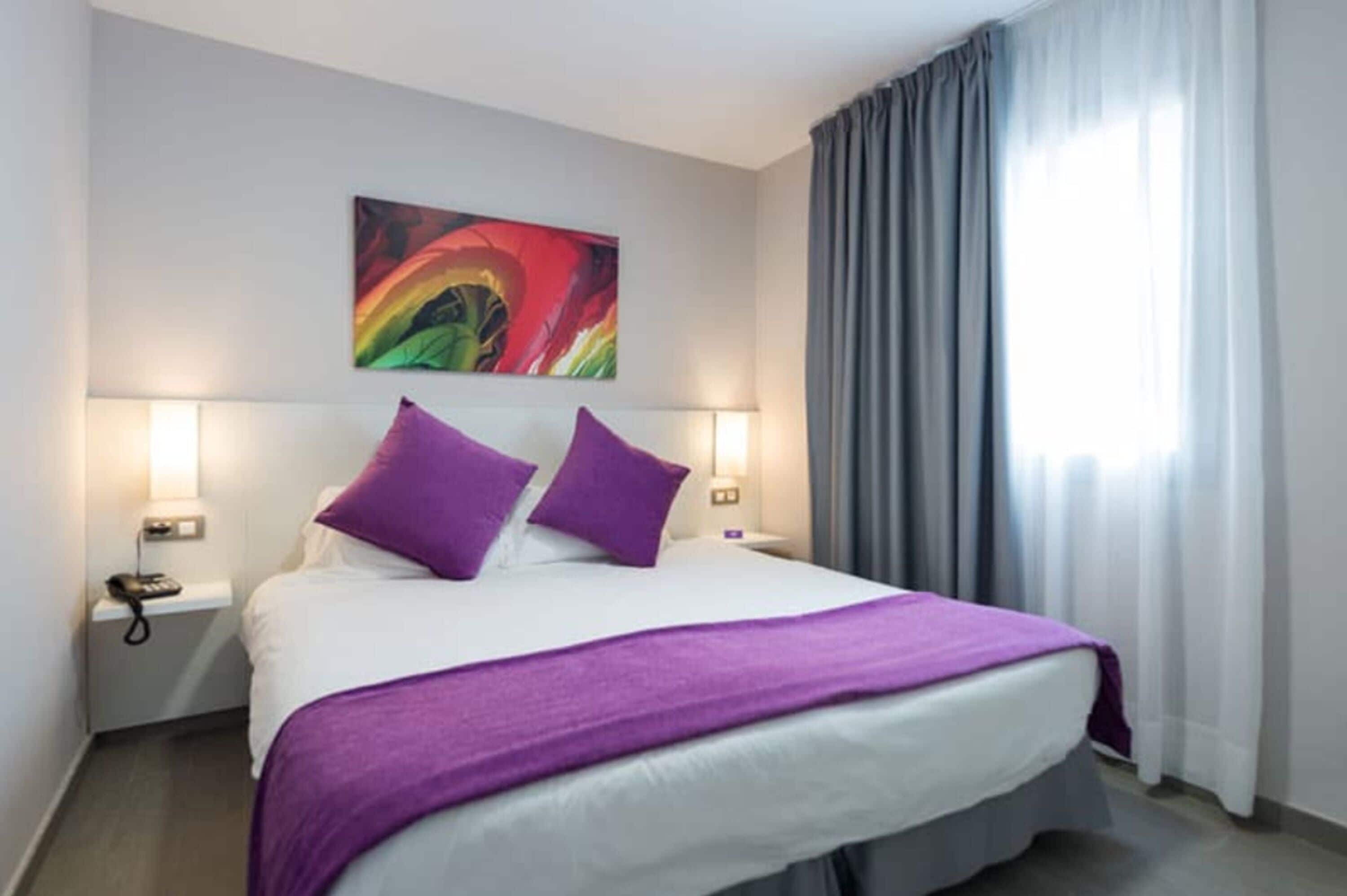 A hotel room with a bed, two purple pillows, a white bedspread, a purple throw blanket, a bedside lamp, a telephone, and a colorful abstract painting on the wall.