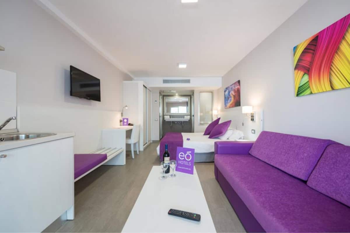 Une chambre d’hôtel moderne équipée d’une kitchenette, d’un canapé violet, d’un lit avec une tête de lit violette et d’un téléviseur fixé au mur.