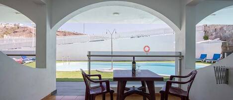 Une terrasse couverte, dotée d’une table et de chaises, qui offre une vue sur une piscine et une zone délimitée par une clôture.