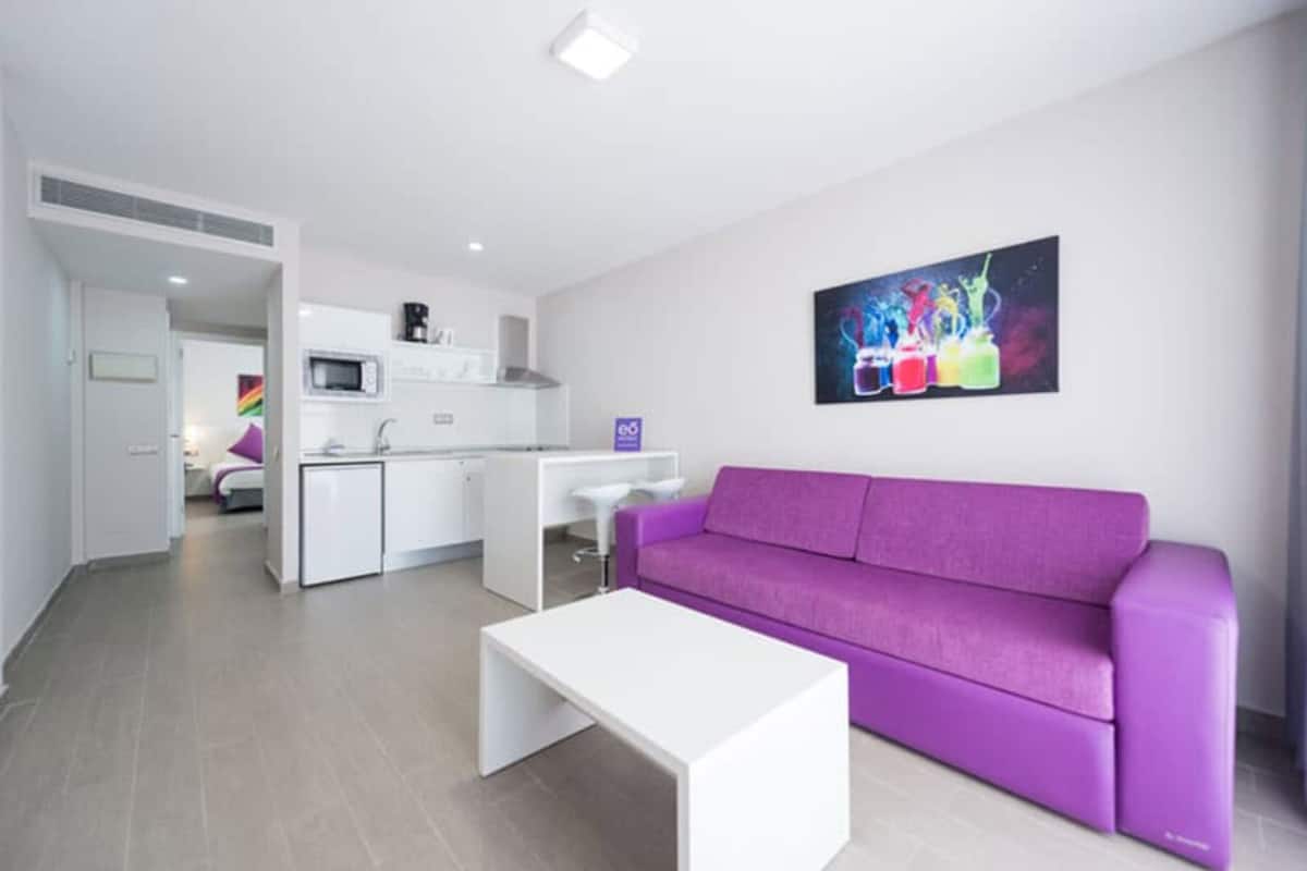 Un salon moderne avec un canapé violet, une table basse blanche et un coin cuisine à l’arrière-plan.