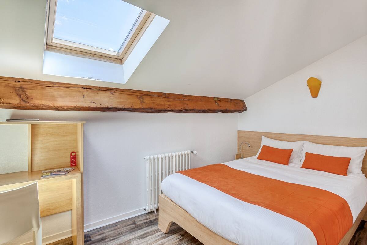 Une chambre avec un puits de lumière, un lit avec une literie orange, une tête de lit en bois, un radiateur et un bureau en bois sur lequel se trouve un magazine.