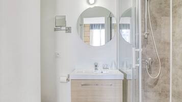 Un baño moderno con lavamanos blanco, espejo redondo, cabina de ducha con mampara de vidrio y pared con azulejos color beige.
