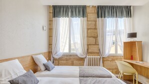 Quarto de hotel com duas camas, uma escrivaninha, uma cadeira e uma janela com cortinas.