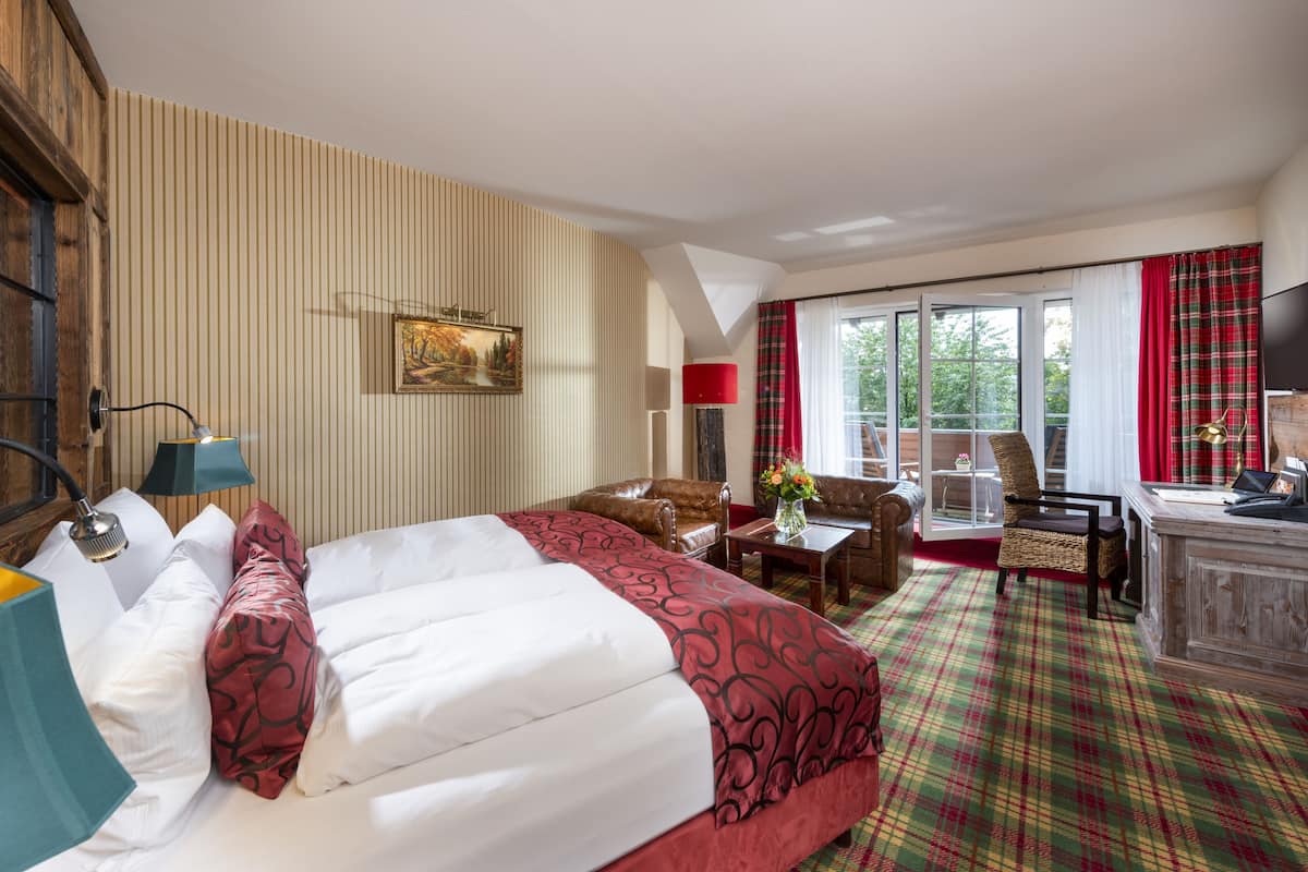 Ein Hotelzimmer mit einem großen Bett, einem Schreibtisch, einem Sessel und einer Sitzecke. Ein Fenster mit Vorhängen bietet Blick auf Bäume.