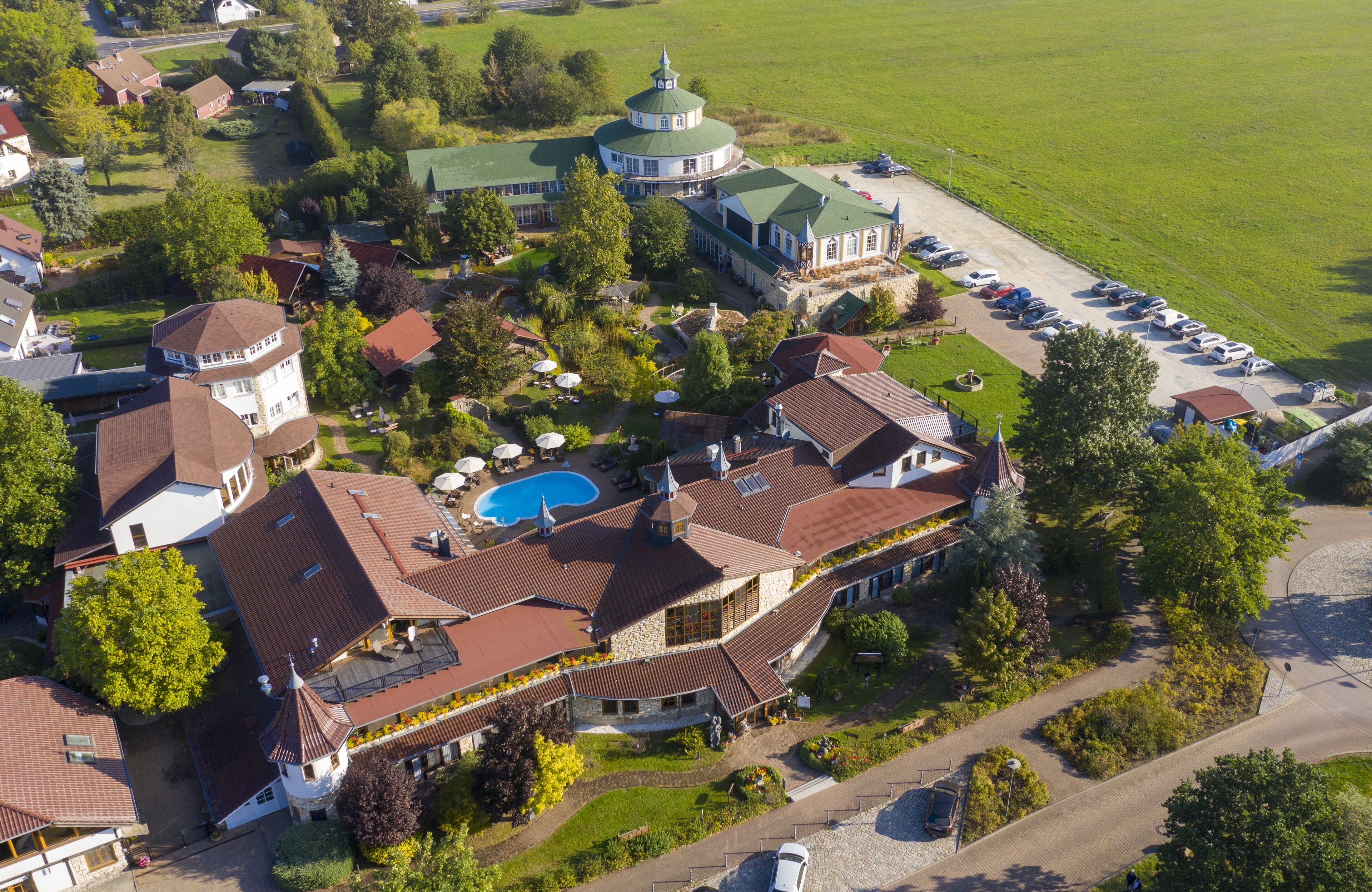 Ein Luftbild eines weitläufigen Anwesens mit mehreren Gebäuden, darunter ein auffälliges Haus mit Swimmingpool, eine Kirche mit Kuppel und ein Parkplatz.