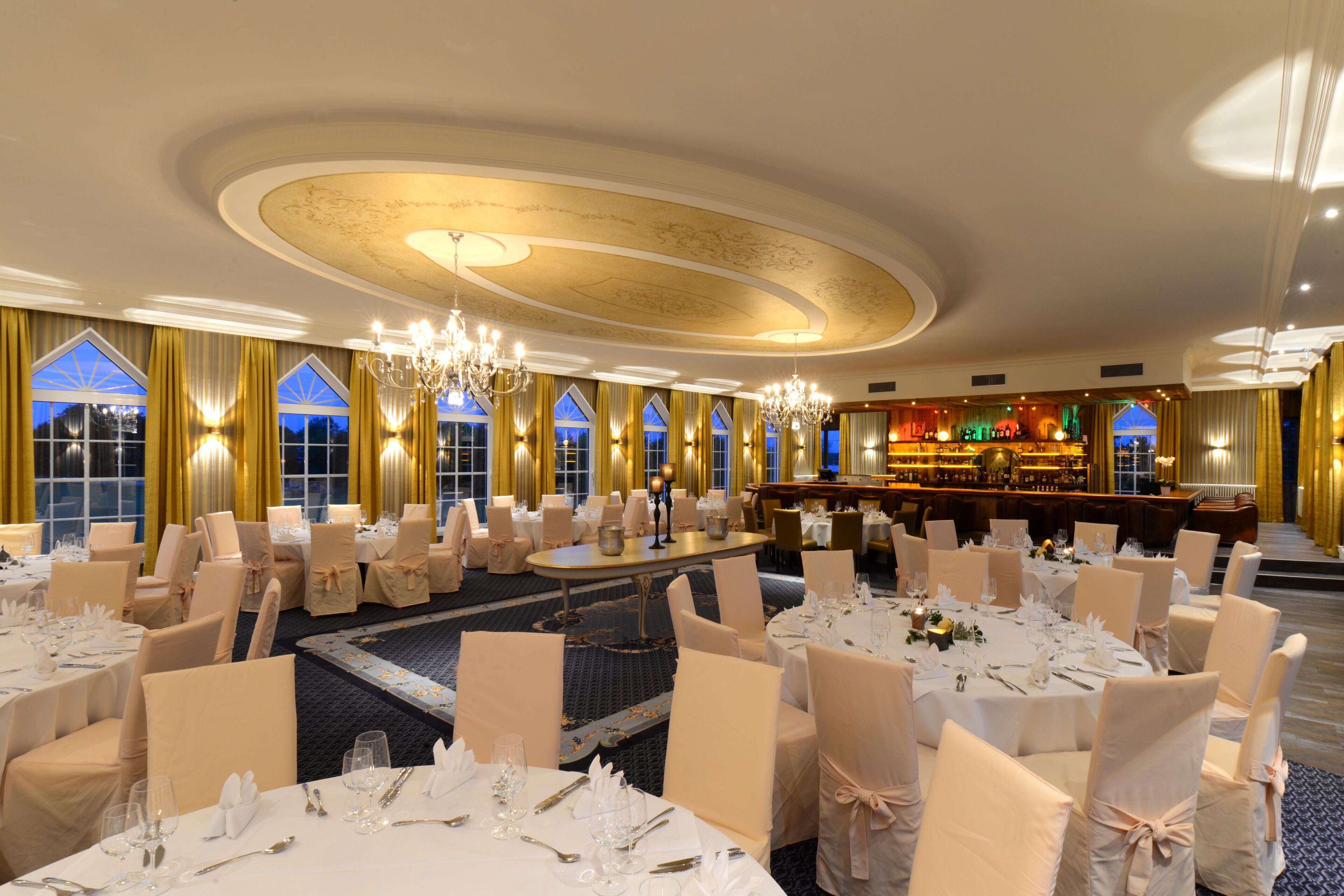 Un salón de banquetes con mesas redondas dispuestas para cenar, una zona de bar y amplias ventanas con cortinas.